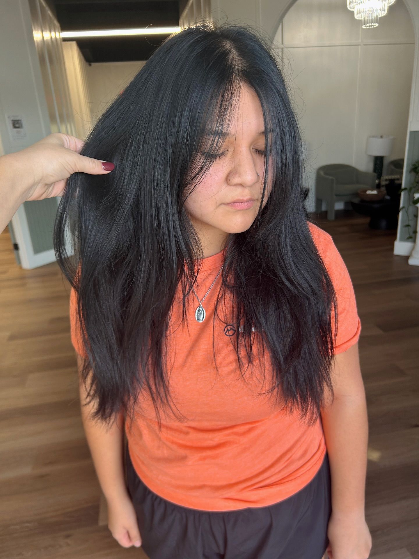 Woman with long, black hair and an orange shirt, eyes closed, in a salon. A hand is holding up her hair.