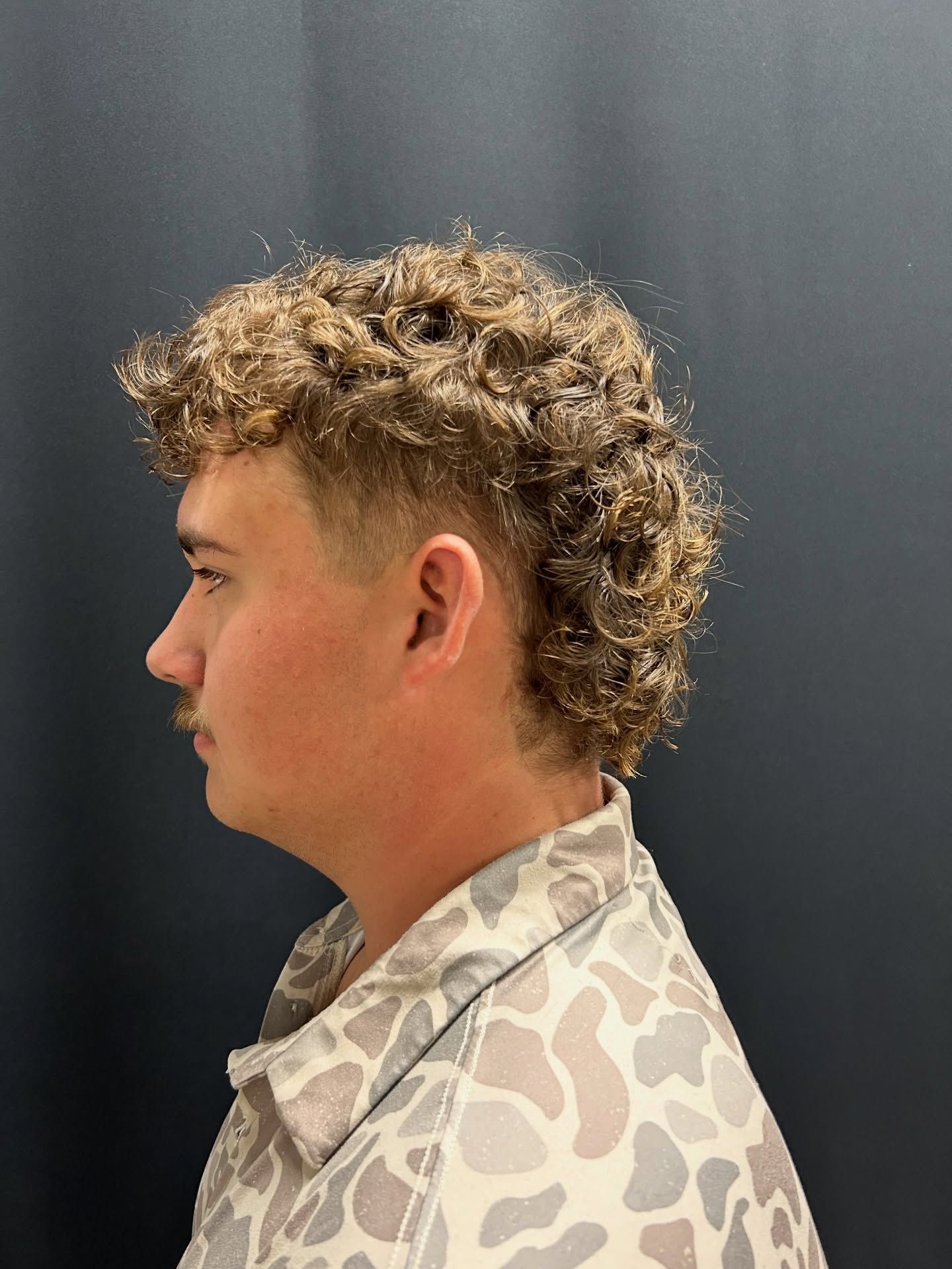Man with curly brown hair in a mullet style, wearing a patterned shirt, against a dark background.