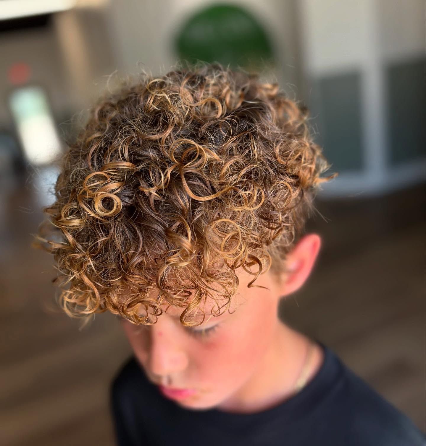 Boy with curly, highlighted hair in a salon setting, looking down.