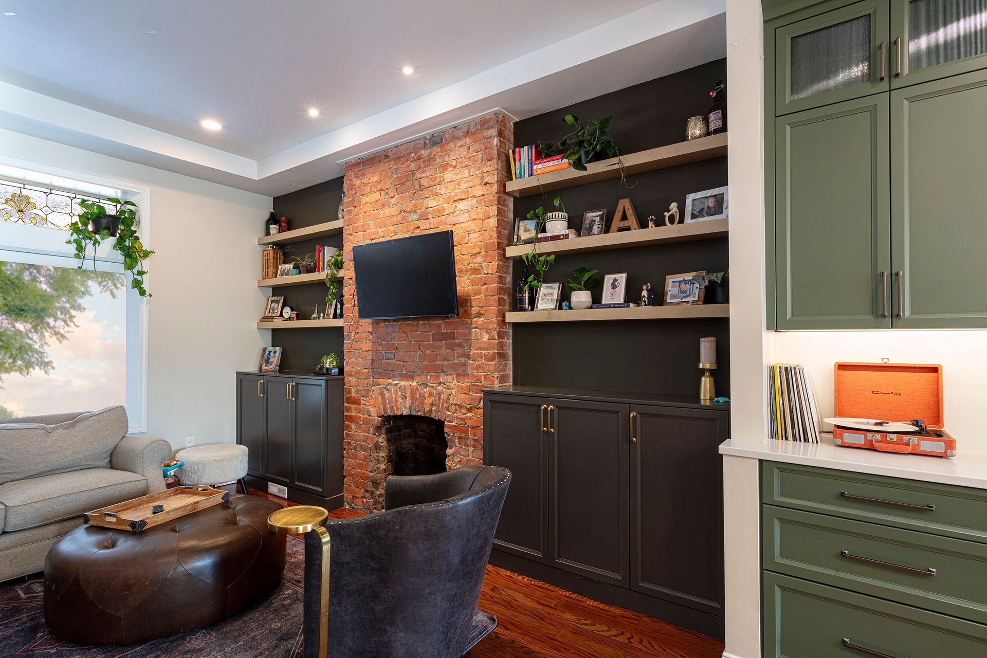 Stylish living room with brick fireplace wall, gray sofa, ottoman, TV, and green built-in cabinets
