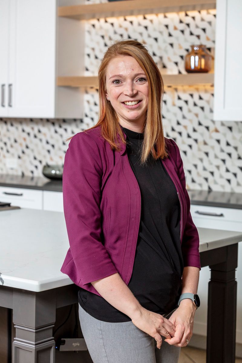 Smiling woman in a purple jacket standing in a modern kitchen with white cabinets and patterned backsplash