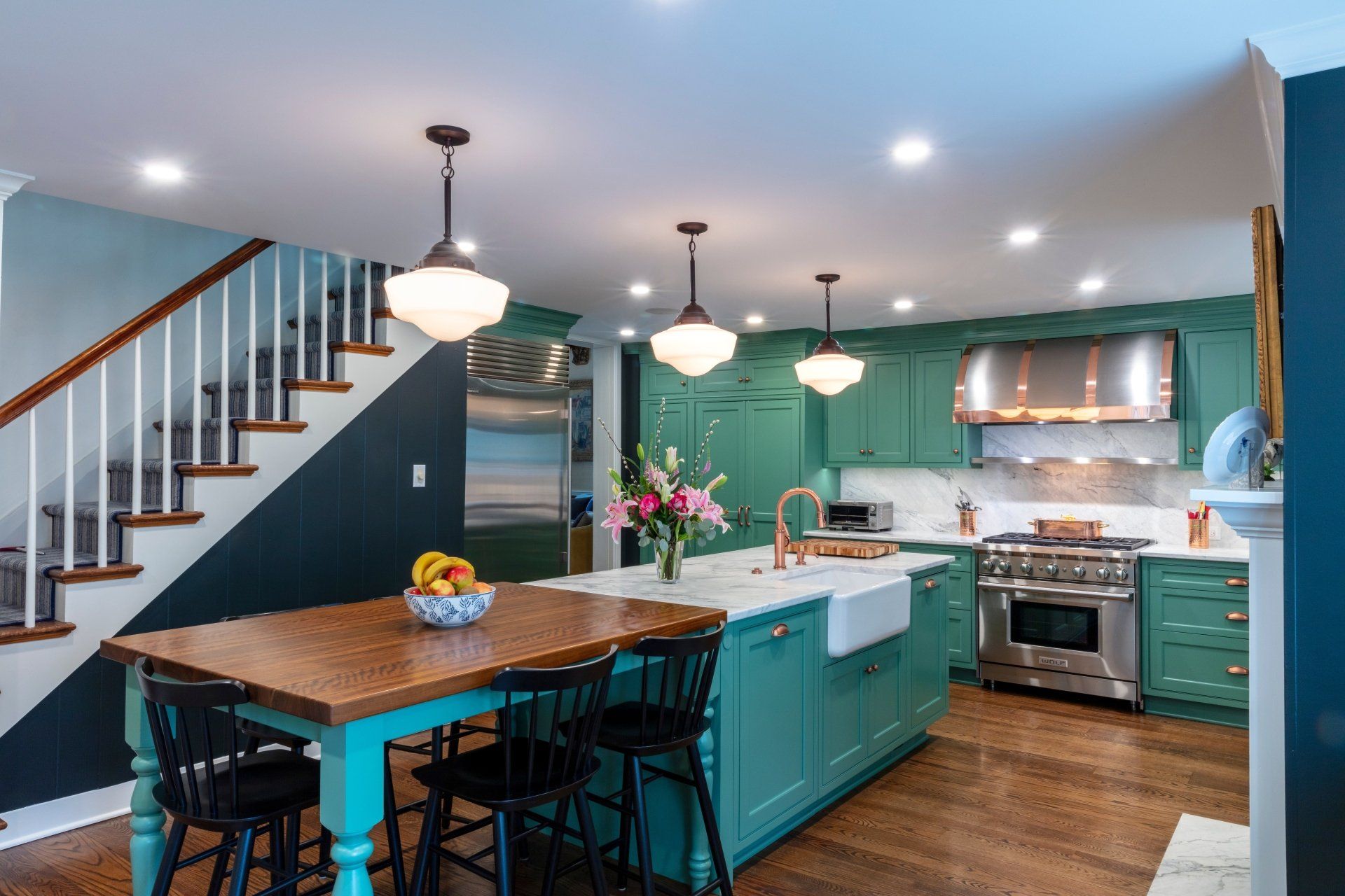 Bright teal kitchen with island, pendant lights, staircase, and wood dining table in an open home layout