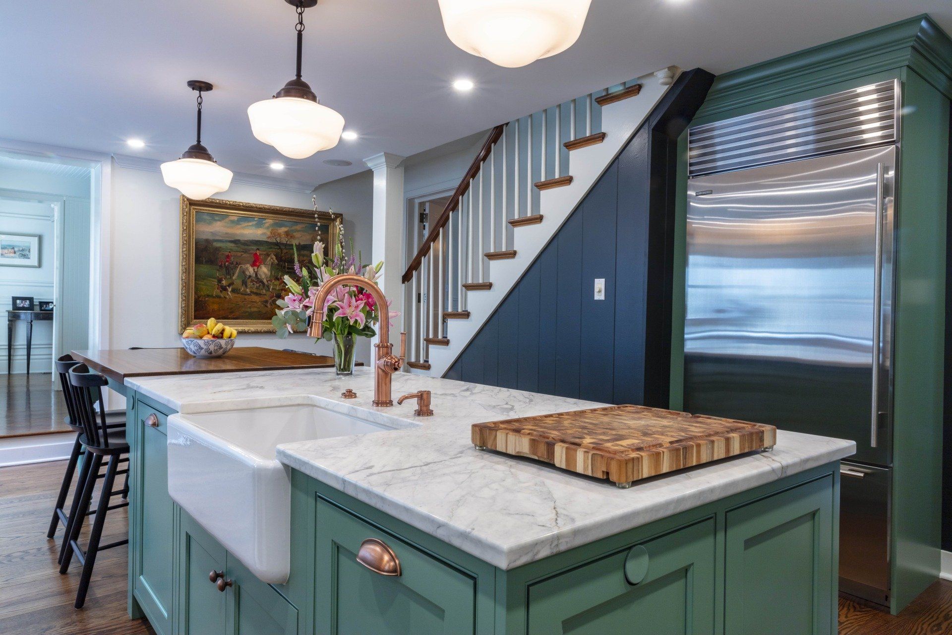 Modern kitchen with teal island, marble countertop, hanging lights, and staircase in the background