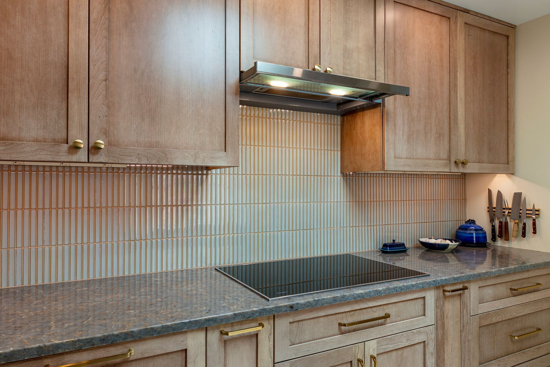Modern kitchen countertop with wood cabinets, gray backsplash, stove, range hood, and blue kettle and teapot
