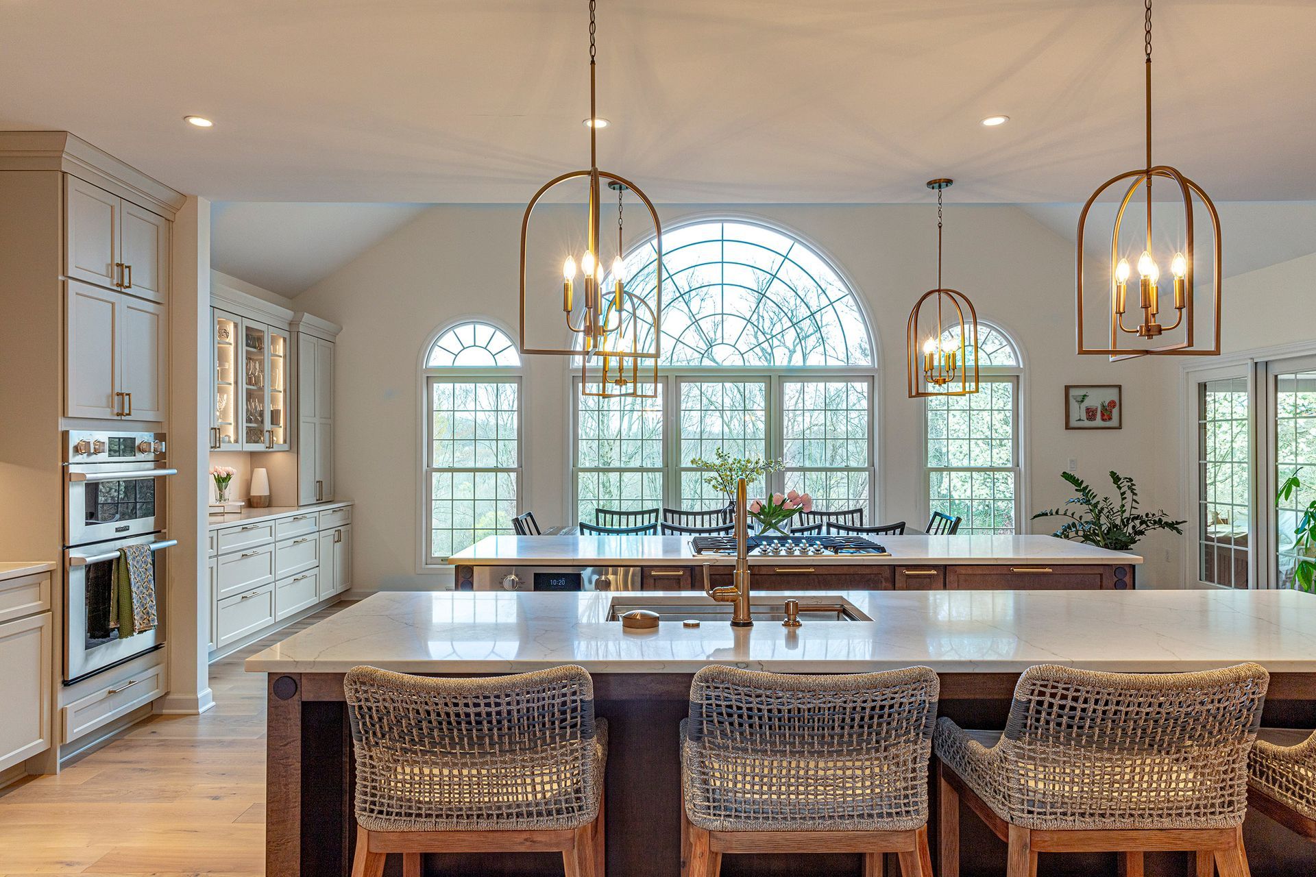 Bright kitchen with a large island, wicker stools, pendant lights, and arched windows overlooking greenery