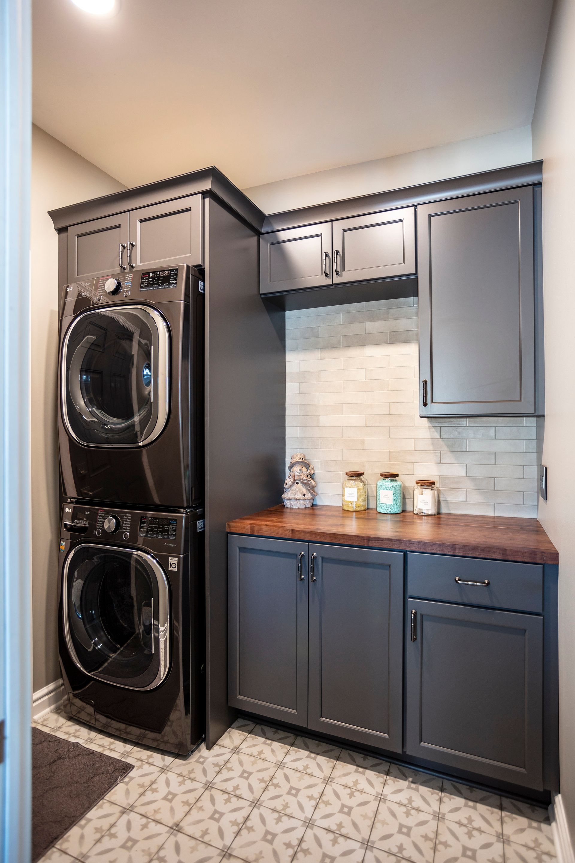 Laundry room with stacked black washer and dryer, gray cabinets, wood countertop, and patterned tile floor