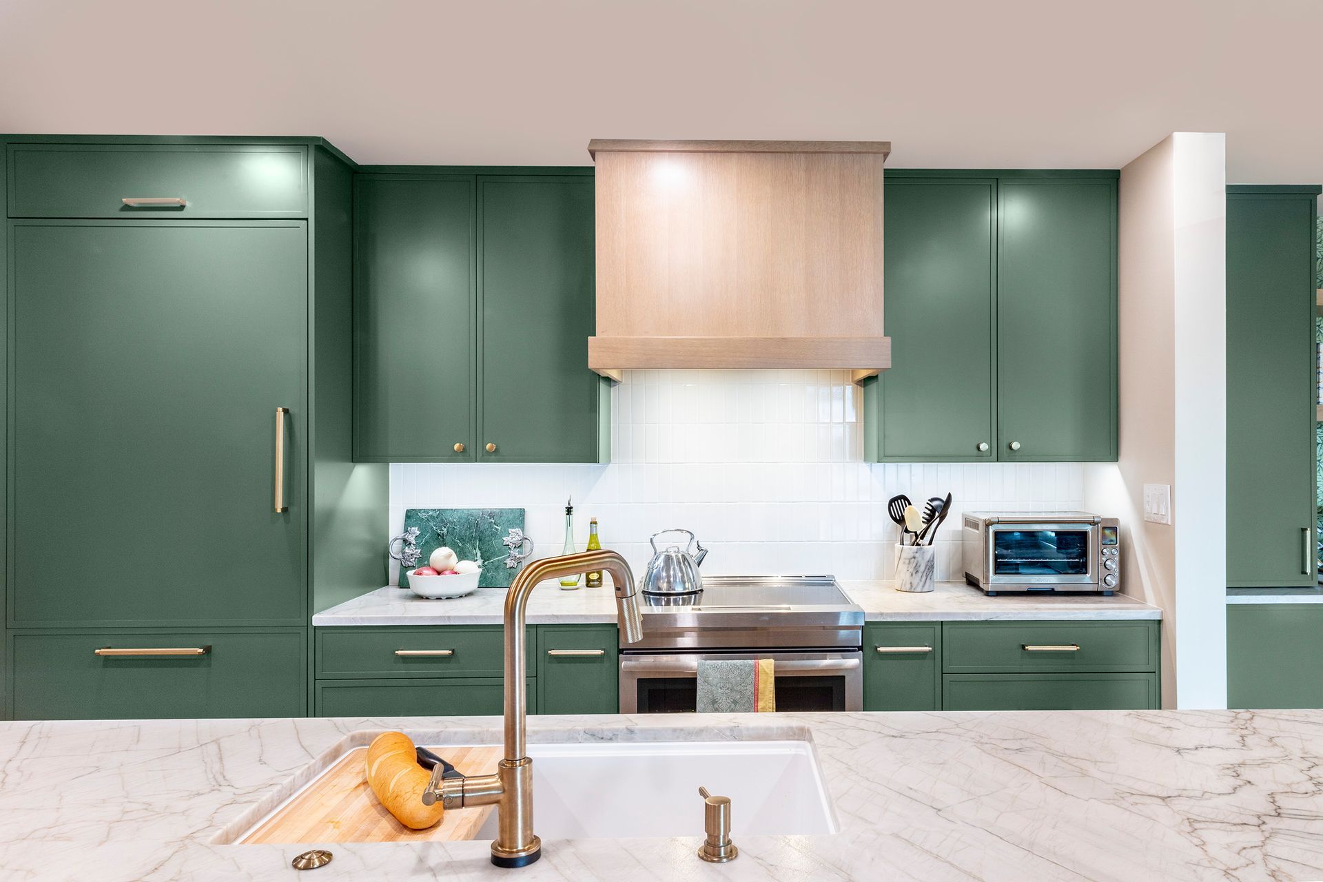 Modern kitchen with dark green cabinets, marble island, brass faucet, and light wood range hood