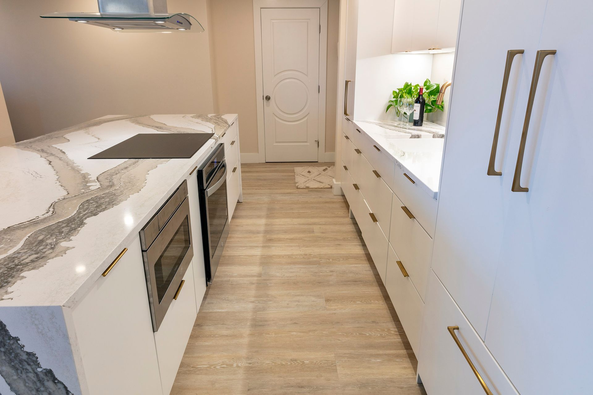 Narrow modern kitchen with white cabinets, marble counters, wood floor, and central door at the end.