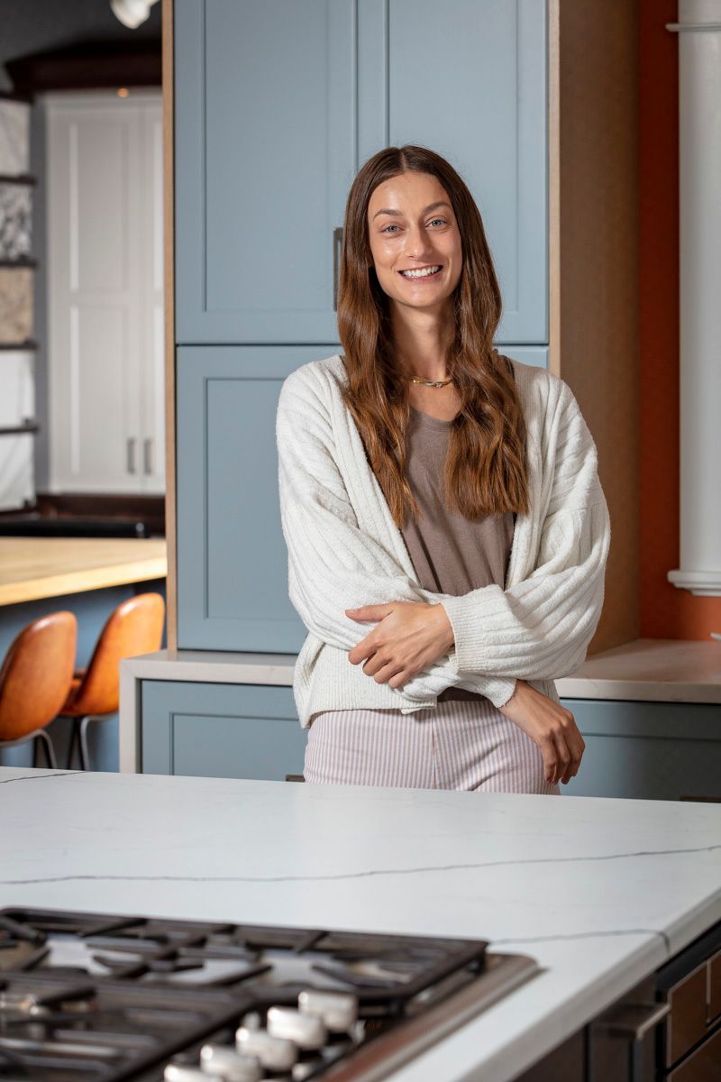 Woman standing in a modern kitchen, smiling with arms crossed, near a countertop and stove.