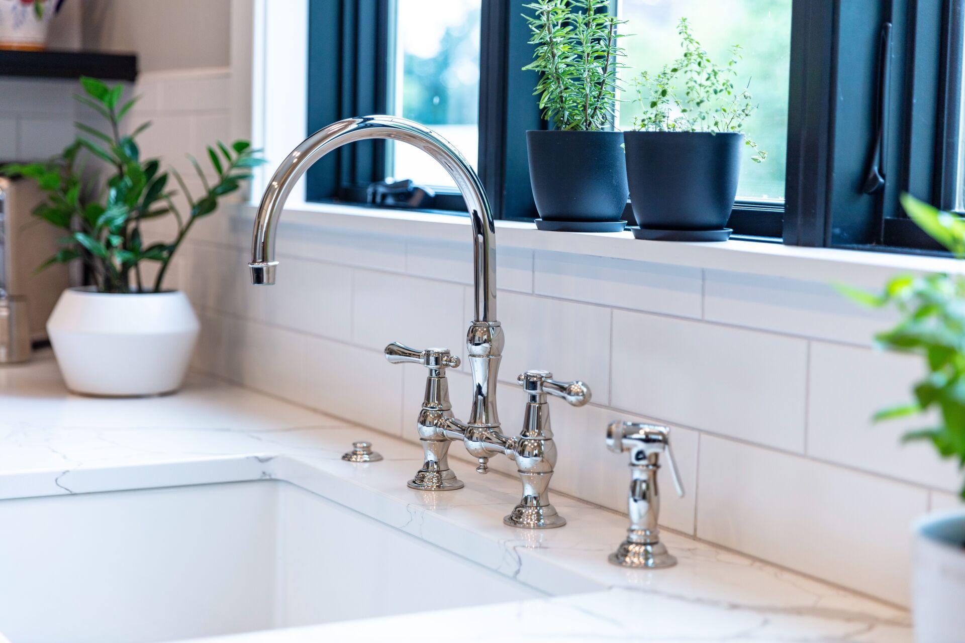 White kitchen sink with chrome faucet beside potted plants on a bright windowsill