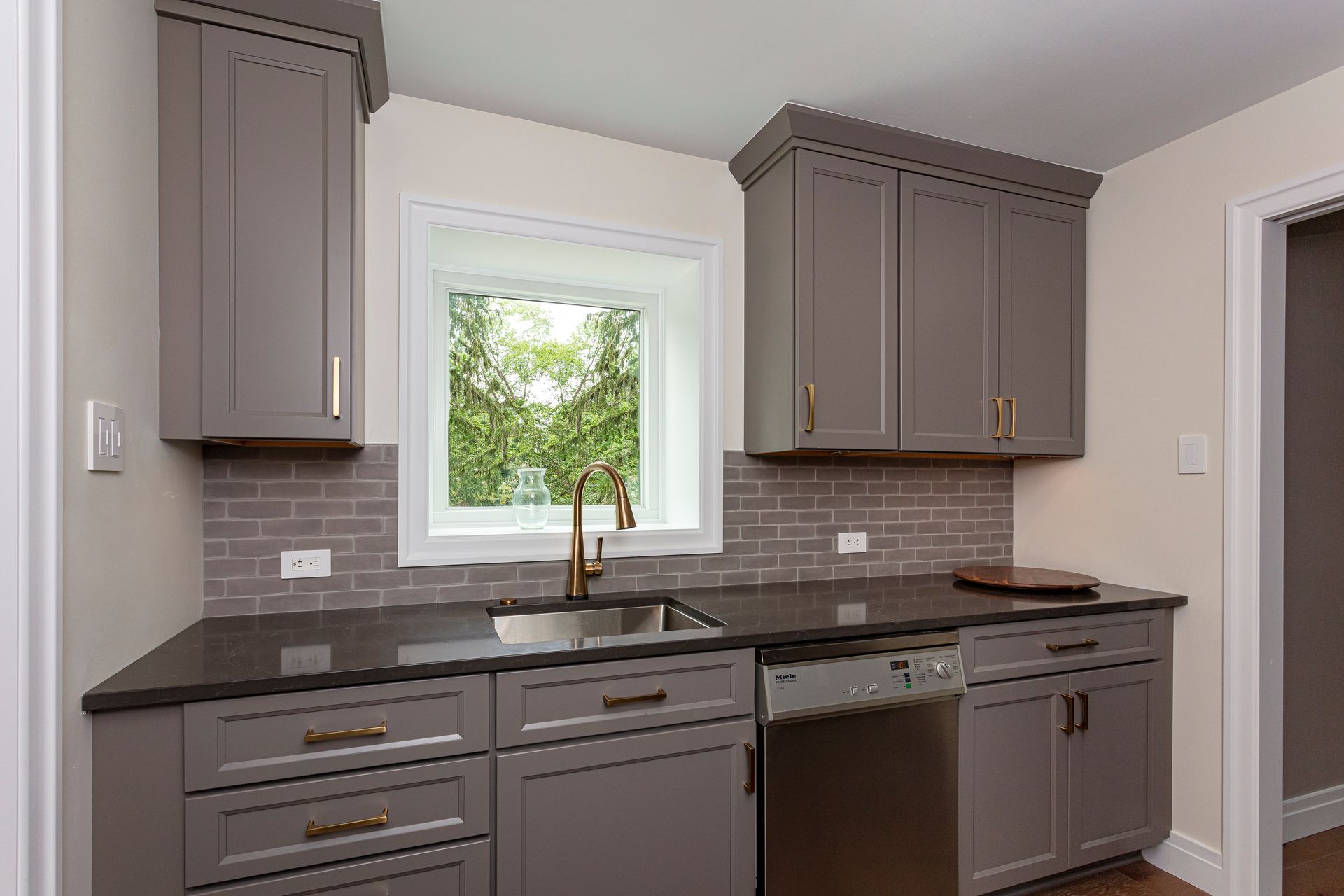 Modern kitchen with gray cabinets, dark countertops, a window over the sink, and a tiled backsplash.