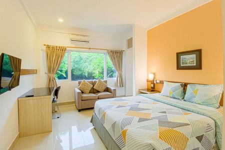 Hotel room with bed, sofa, desk, and TV; window with curtains and foliage view.