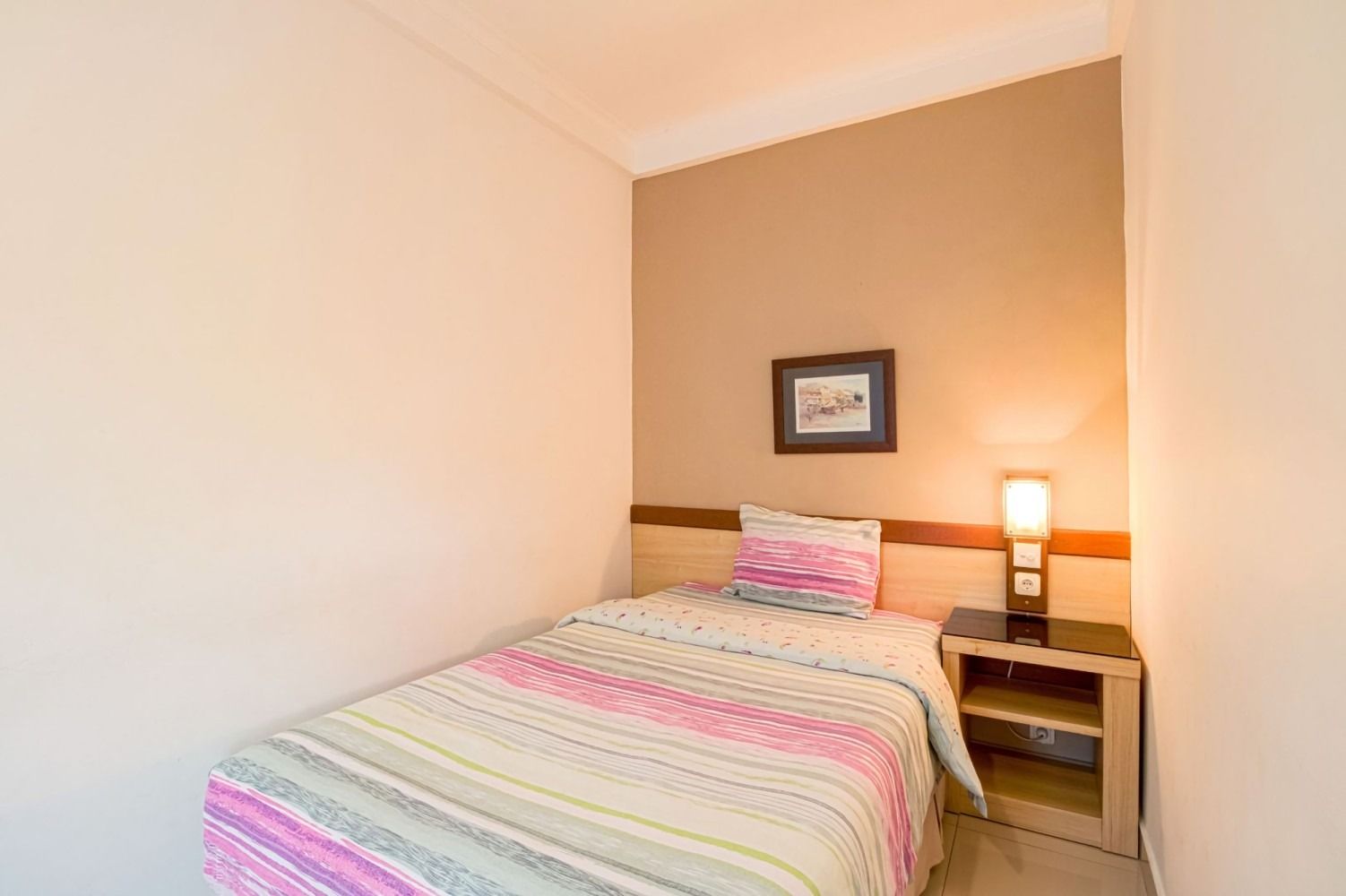 A small hotel room with a single bed and a nightstand. The walls are light beige and the bed has pink and white stripes.
