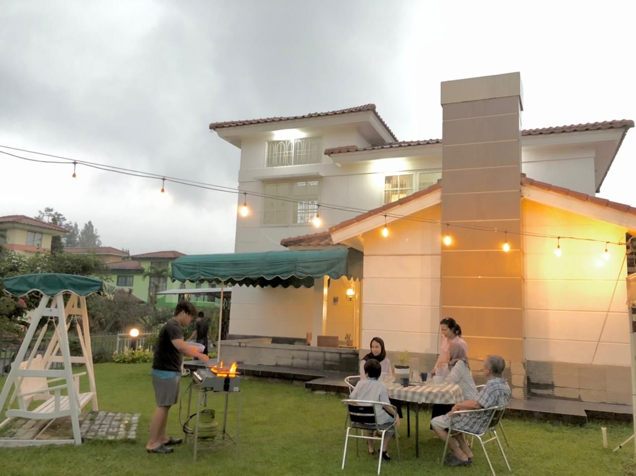 People gathered for a barbecue in a backyard, near a white house, under string lights.