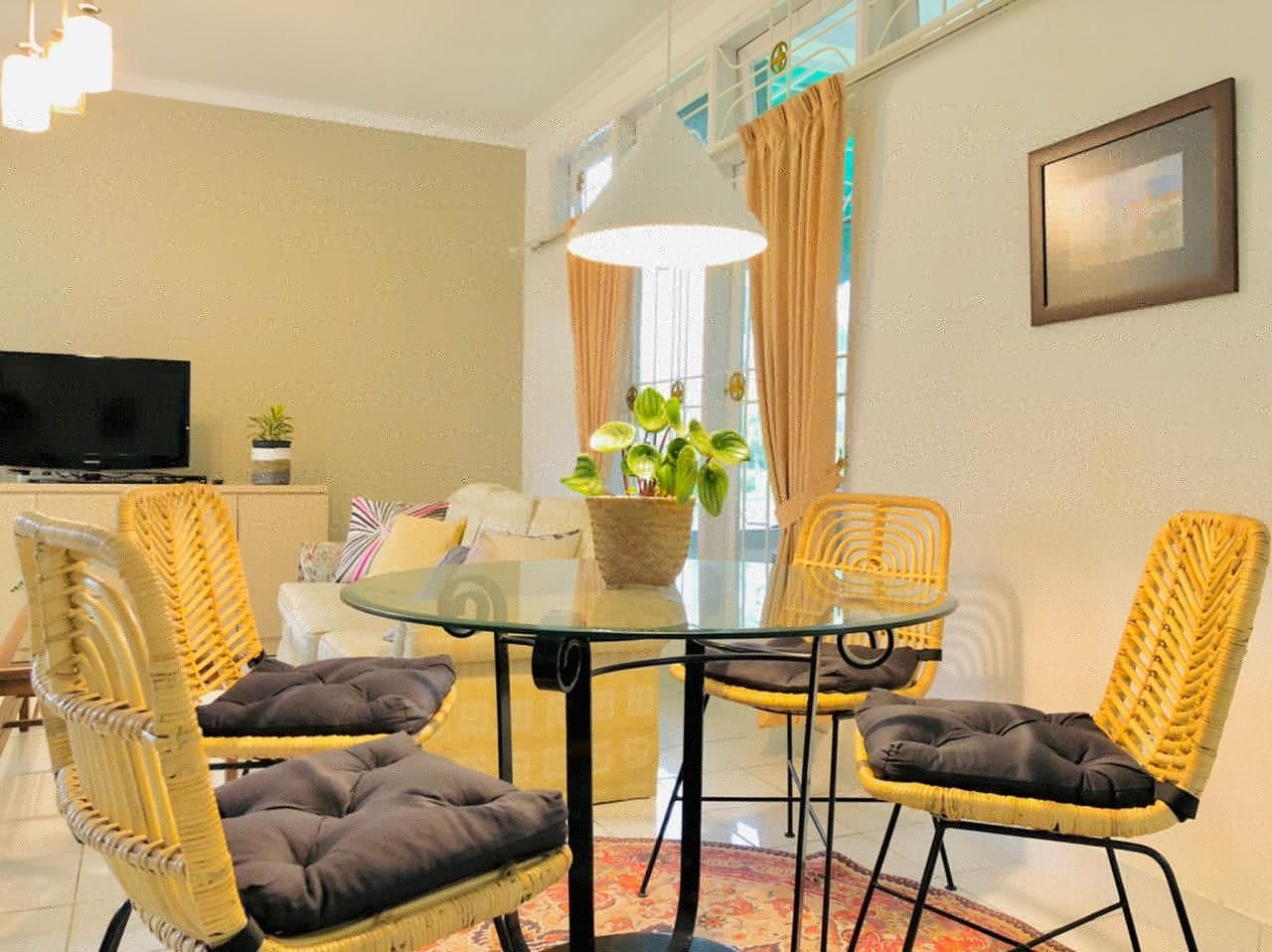 Dining area with glass table, woven chairs with black cushions, and a plant. Beige walls, window, TV in background.