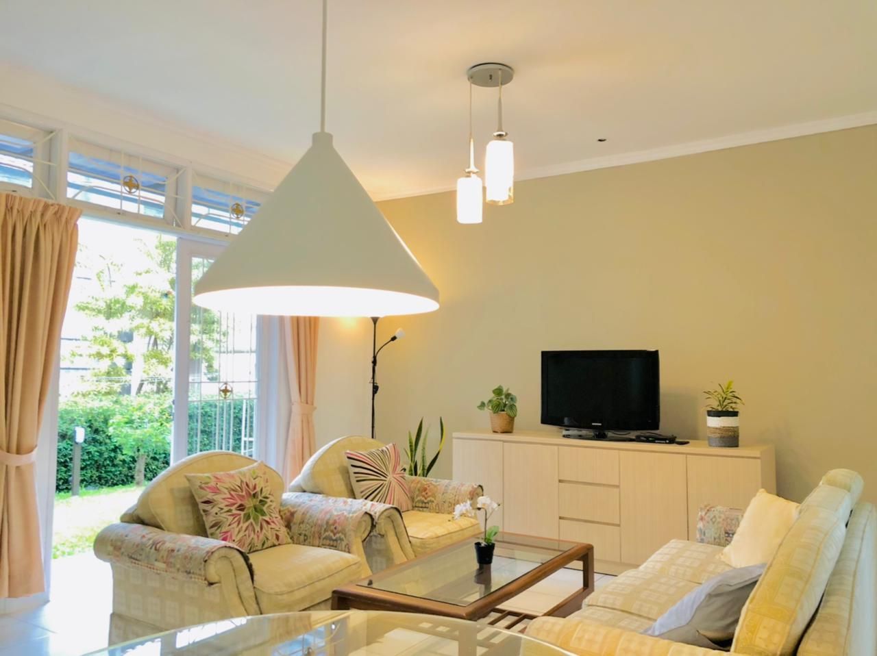 Cozy living room with cream-colored sofas, a TV, and large windows overlooking a green yard; modern pendant lights.