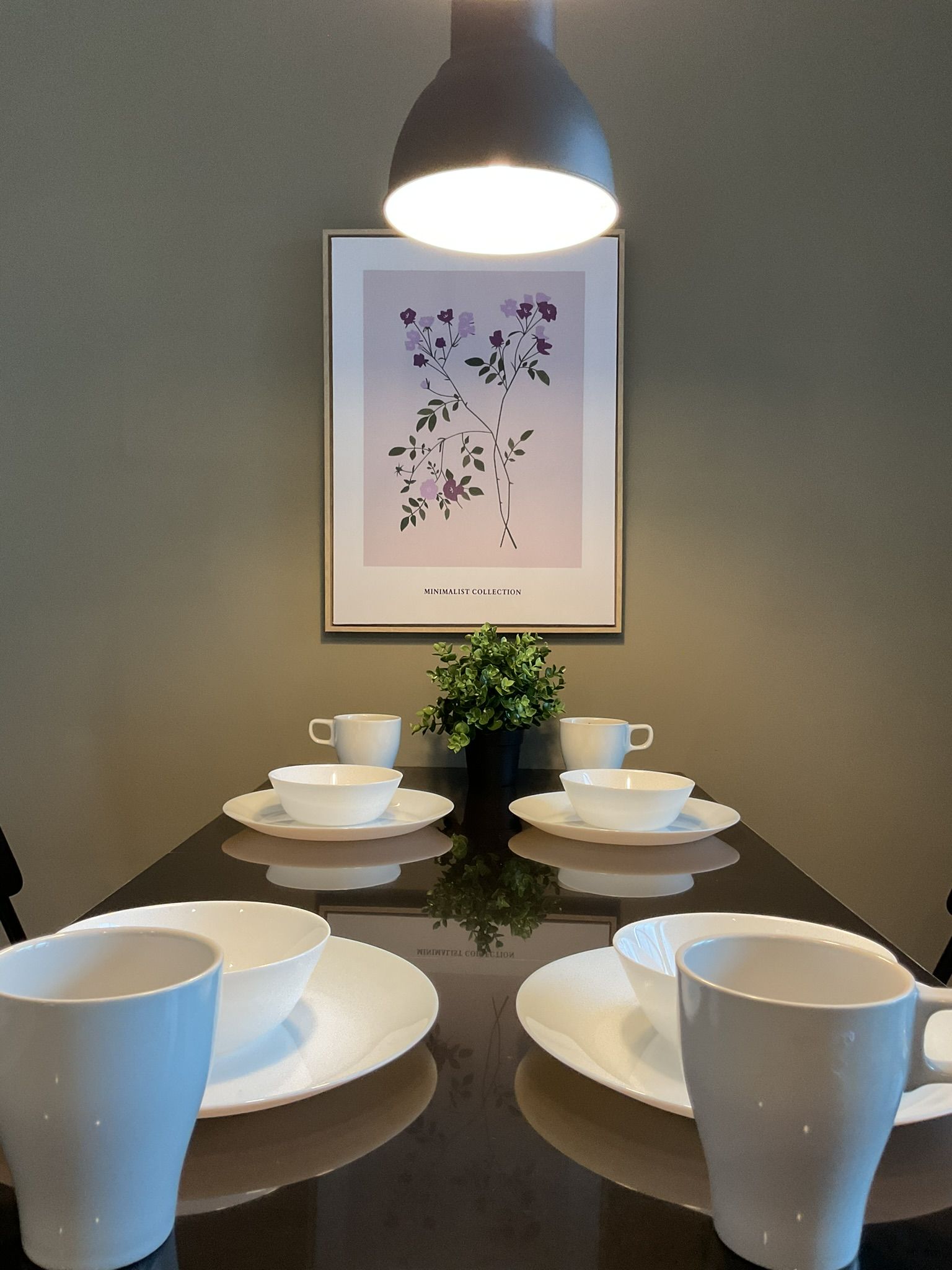 Dining table set for four under a hanging light, with artwork and a small plant.