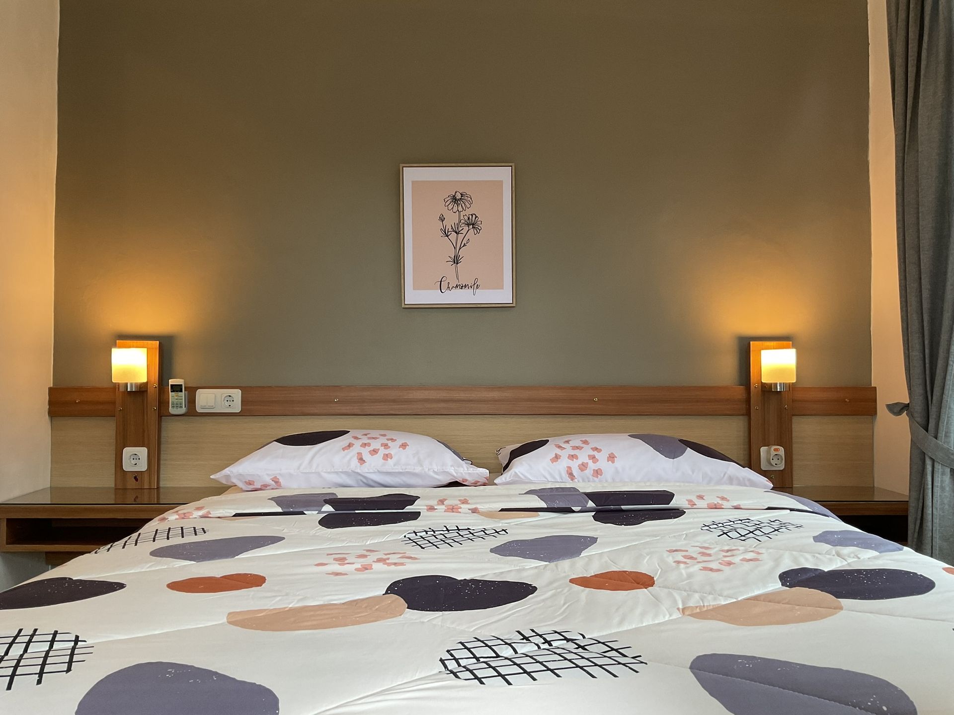 Bedroom with bed, patterned comforter, lamps, framed art, and neutral-colored wall.