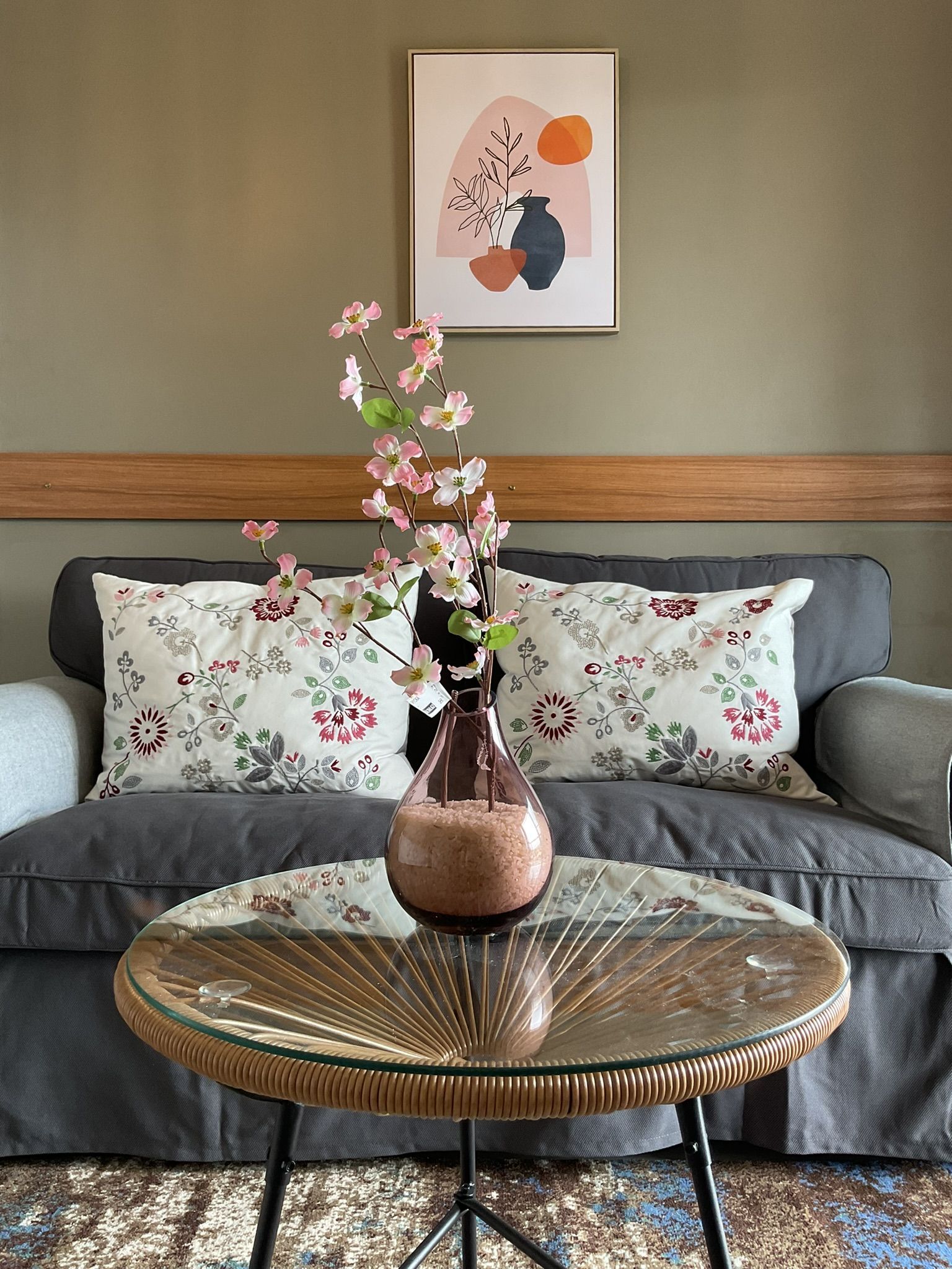 A living room with a gray sofa, a brown coffee table with a vase of pink flowers, and a framed abstract art.