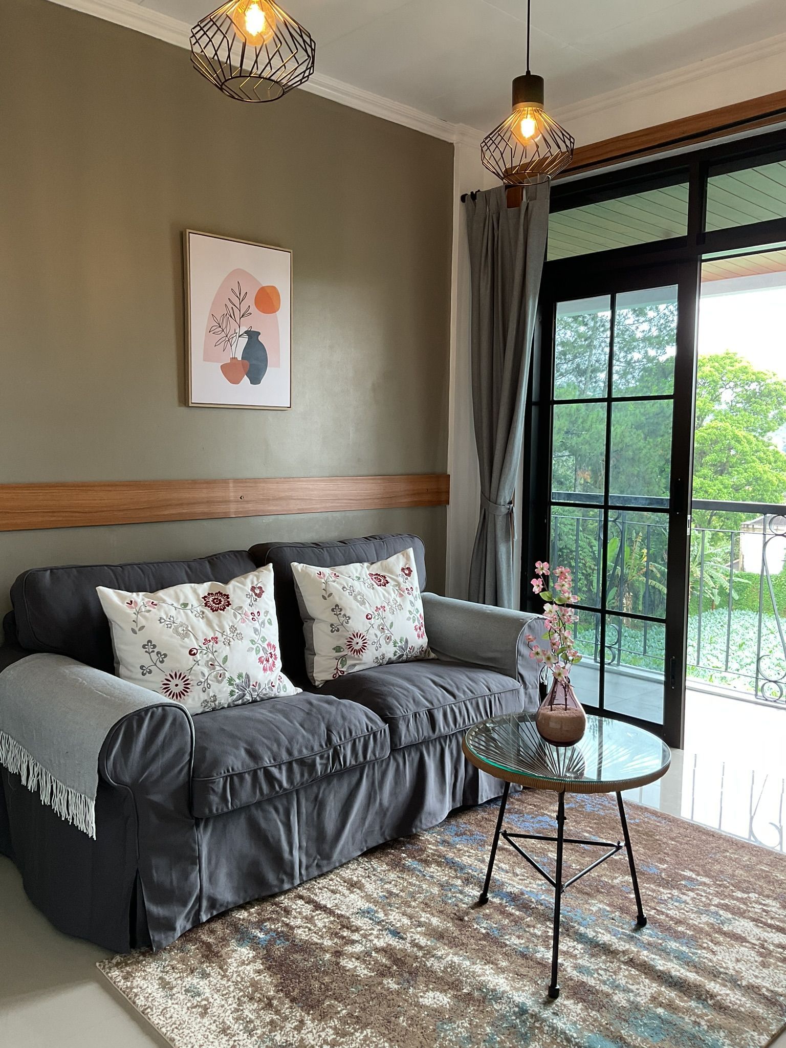 Cozy living room with a gray sofa, floral pillows, and a small glass table.  Artwork on a green wall, and balcony access.