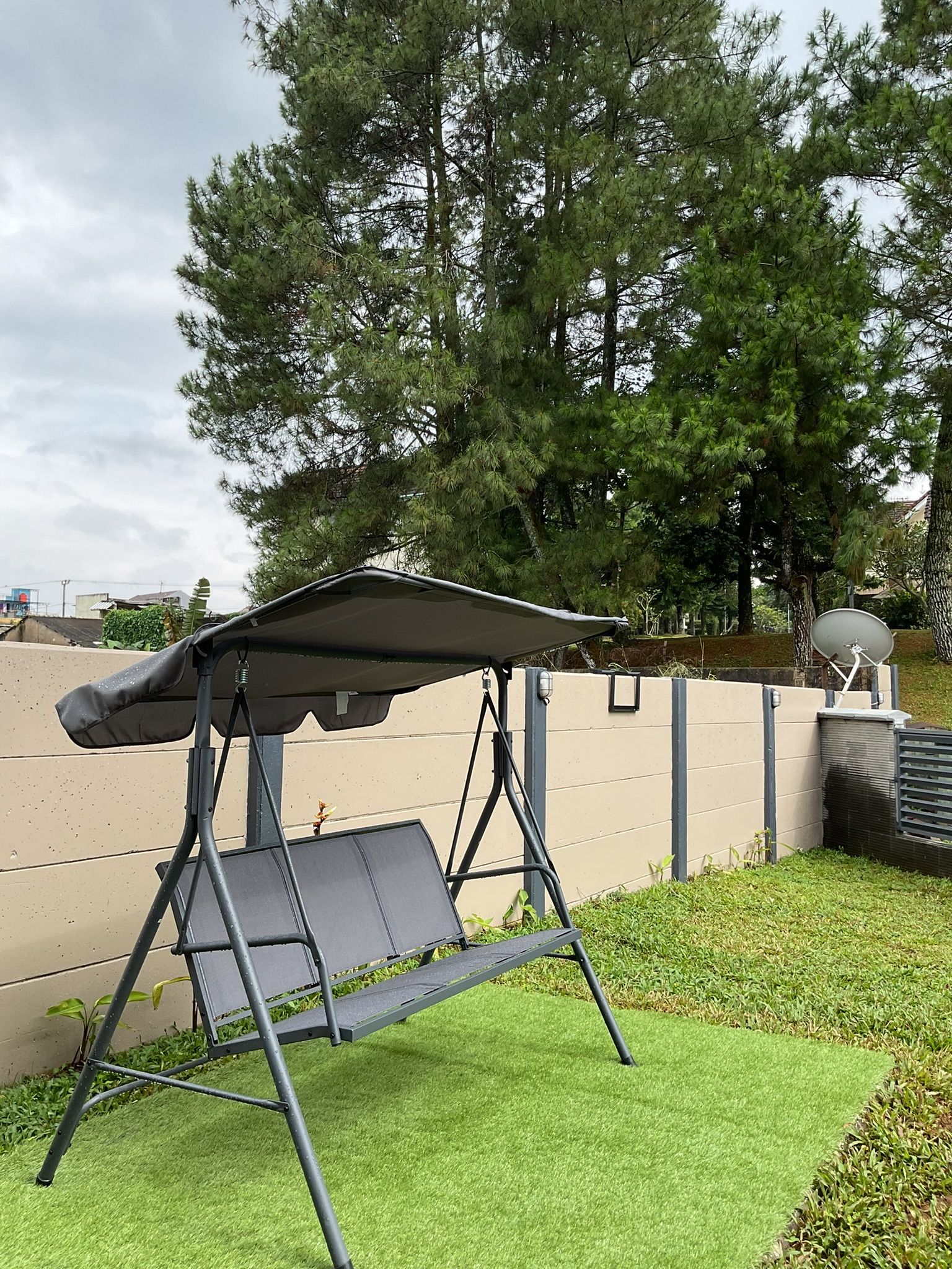Gray swing set with canopy on artificial grass, in front of a tan fence and trees.