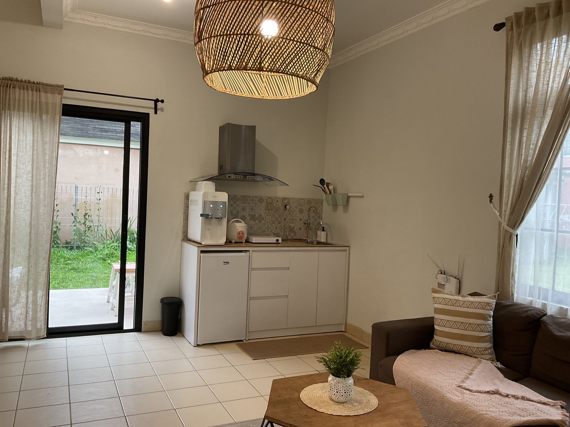 Interior shot of a small kitchen area with a sofa, opening onto a garden. Neutral colors, natural light.