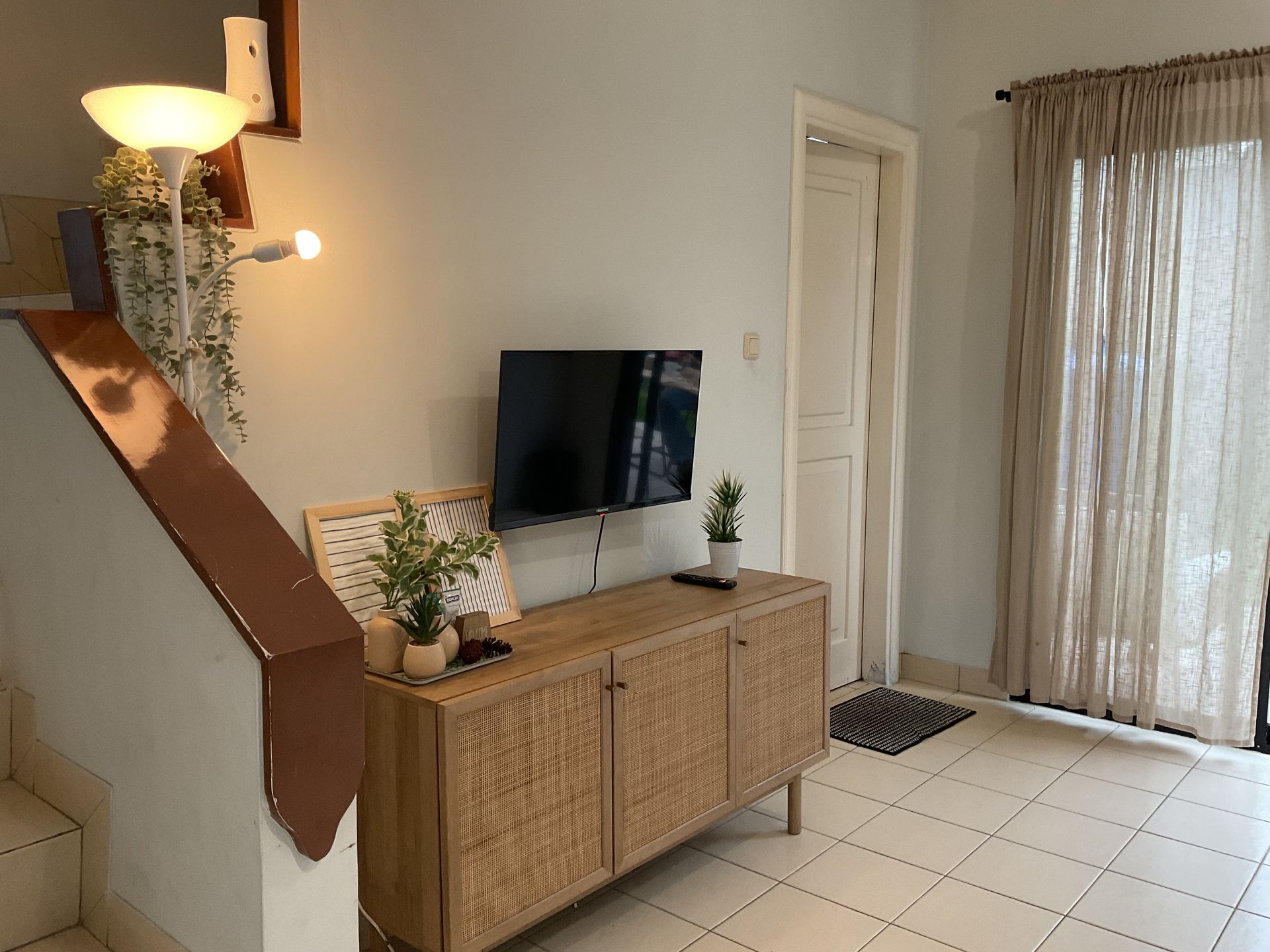 Living room with TV on woven cabinet, next to stairs and window with curtains.