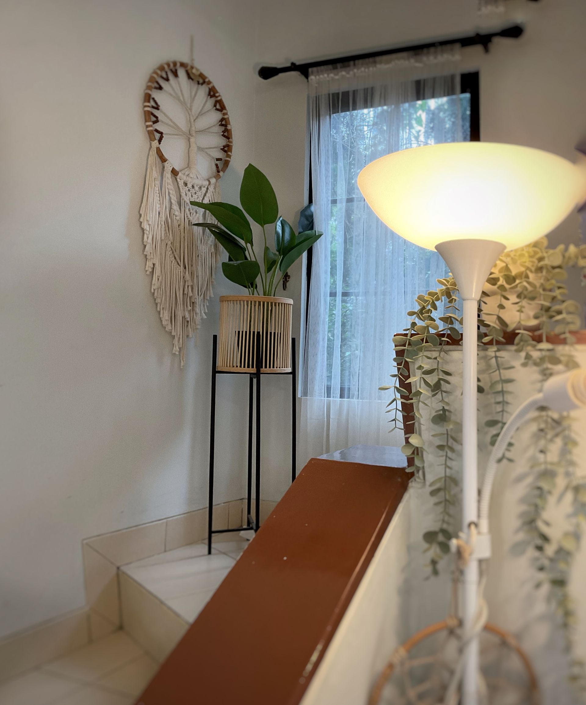 Stairwell with dreamcatcher, potted plant, and floor lamp. Window with sheer curtain in the background.