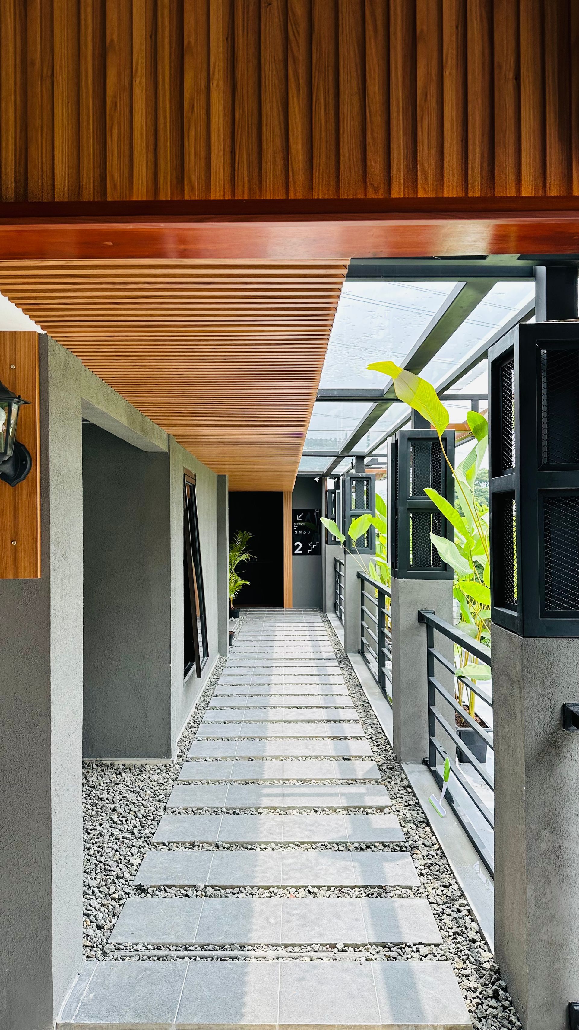 Long hallway with stone pavers and pebbles, lined with a wooden ceiling and an open-air balcony with greenery.