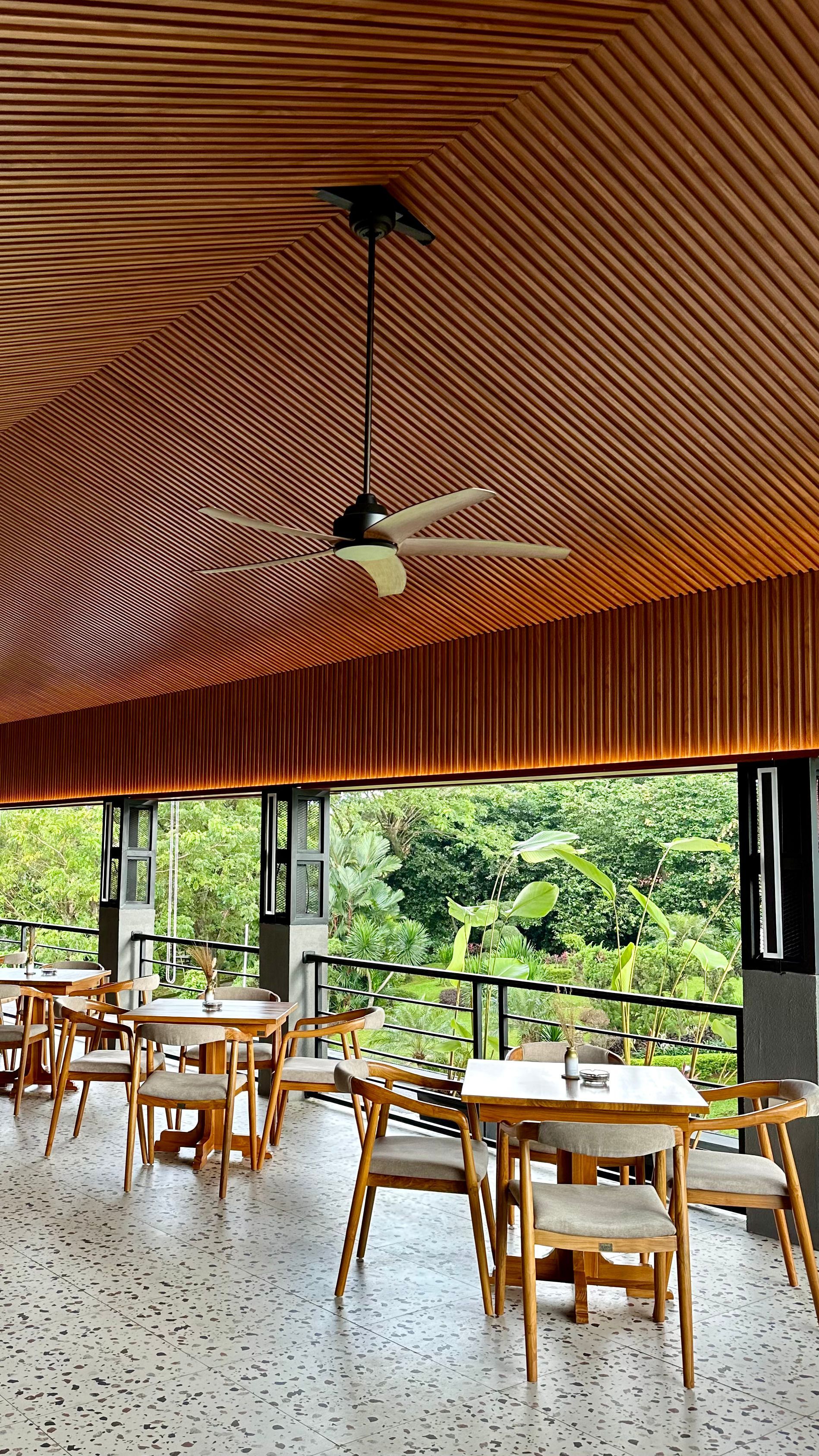 Restaurant interior with wooden ceiling, tables, chairs, and a view of lush greenery.