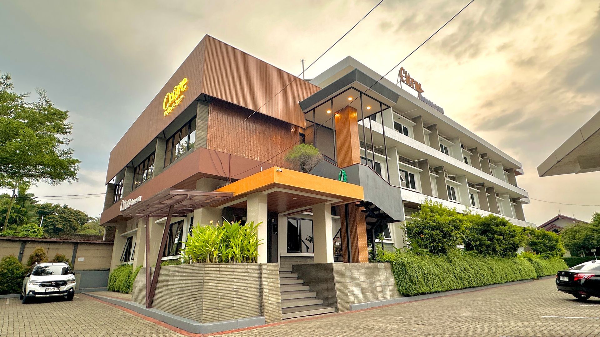 Hotel exterior with brown, orange, and gray building, featuring signage, plants, and parked cars.