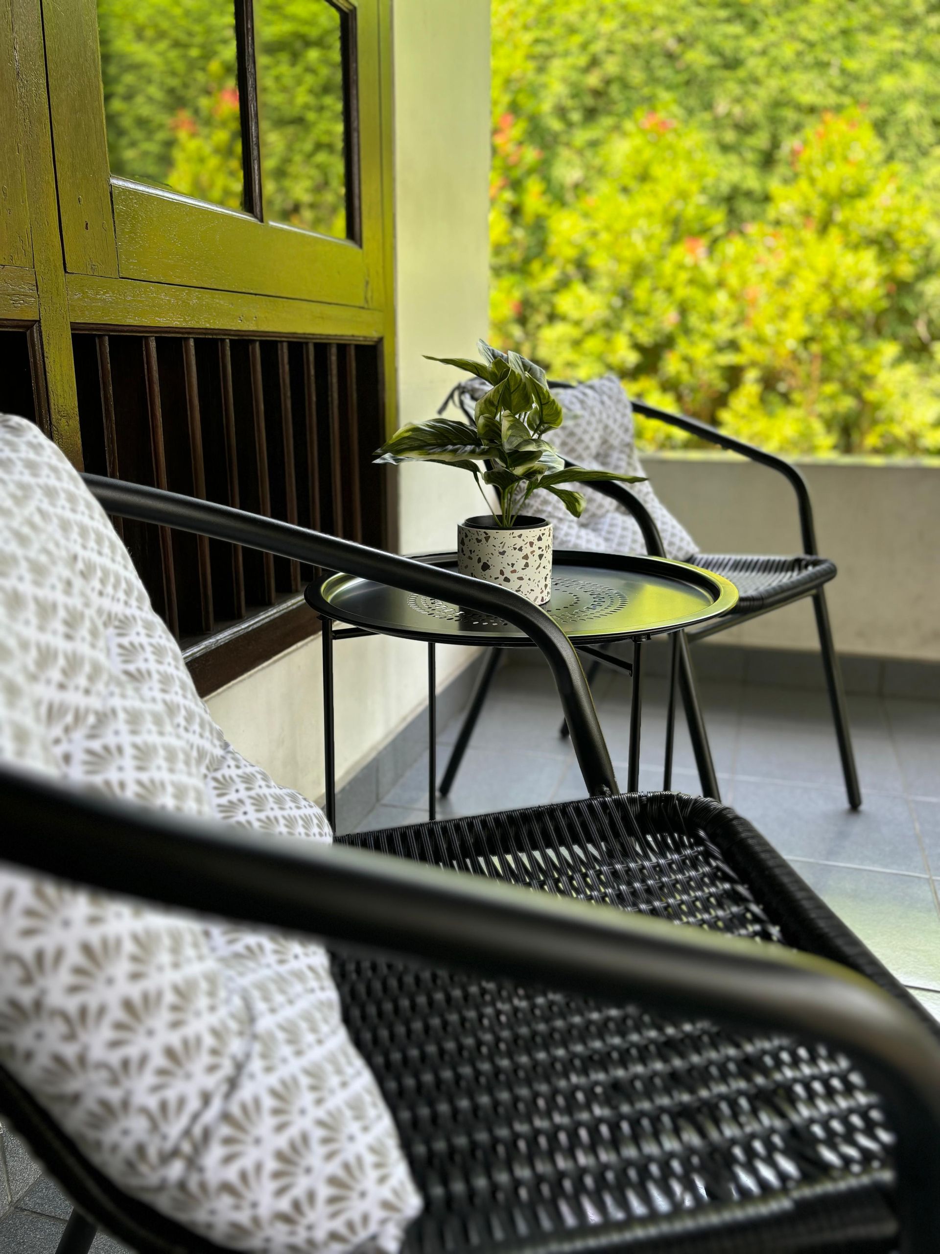 Black wicker chairs and a small table on a balcony overlook lush greenery; a potted plant sits on the table.