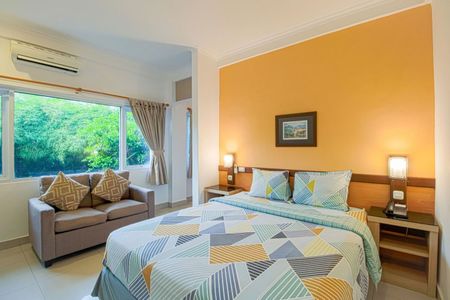 Bedroom with bed, sofa, and window overlooking trees. Orange wall with art, neutral colors.