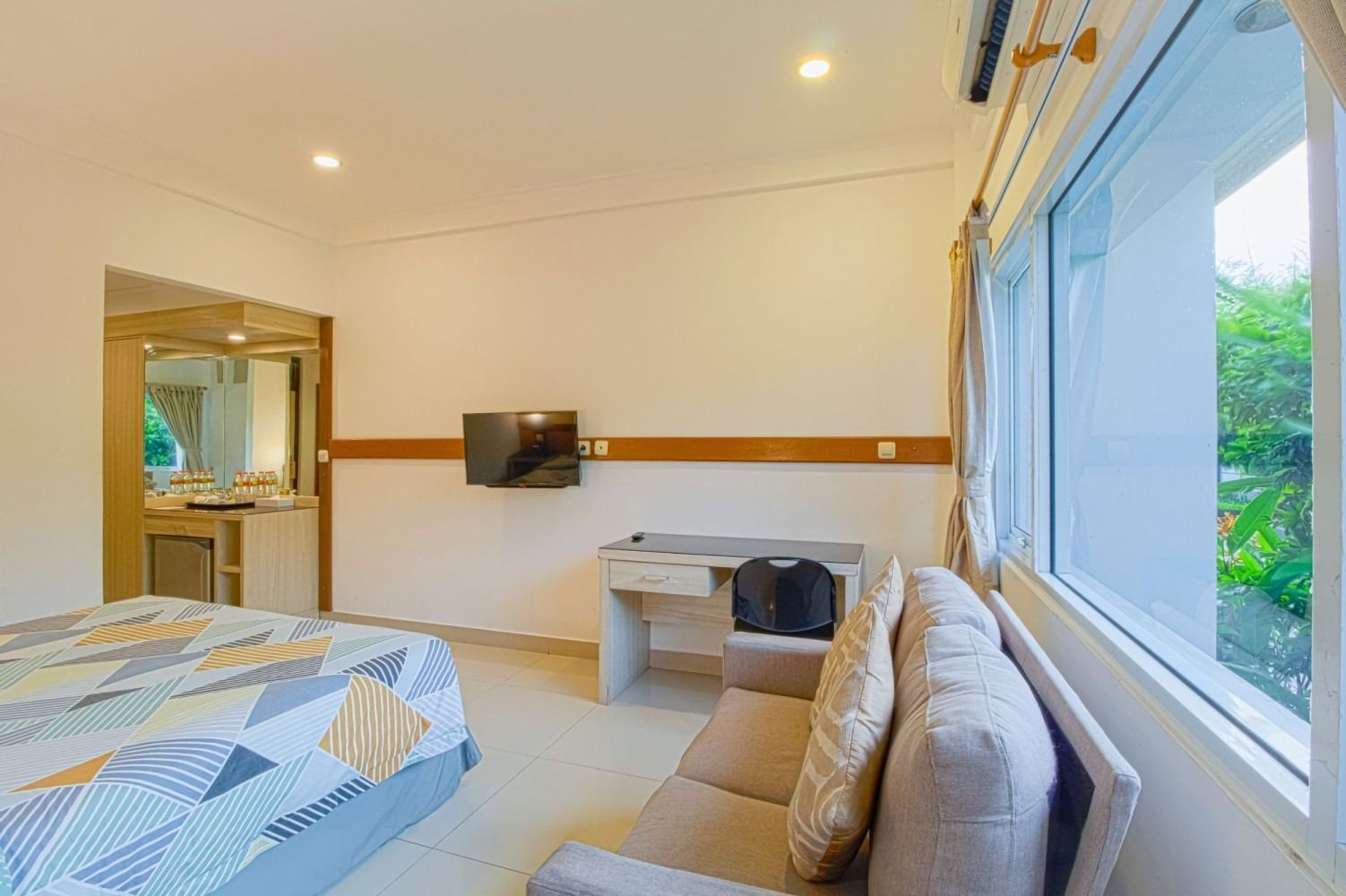 Hotel room with bed, sofa, desk, TV, and large window overlooking greenery.