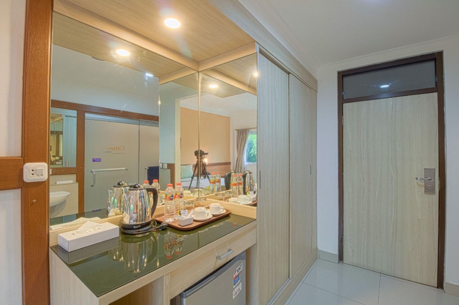 Hotel room interior: vanity with mirror, electric kettle, mini-fridge, and wardrobe next to a closed door.
