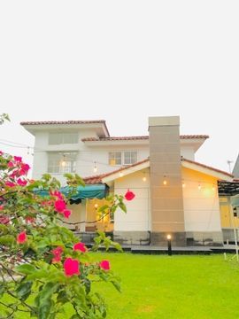 White two-story building with a red-tiled roof, green lawn, pink flowers in the foreground, and string lights.