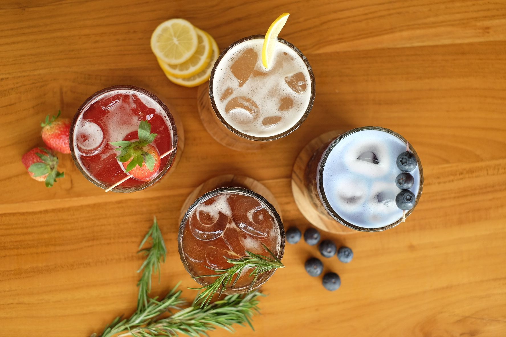 Four colorful cocktails on a wooden surface, each garnished with fruit and herbs.