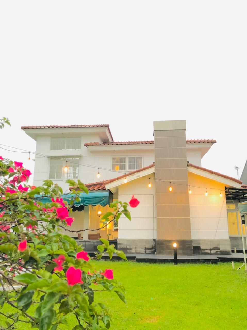 White two-story house with red tile roof, chimney, green lawn, and pink flowers in foreground. String lights.