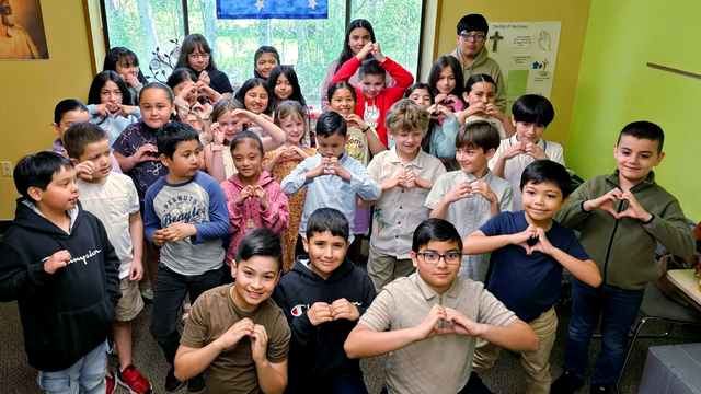 A group of children in a classroom posing together, smiling and making heart hand gestures.