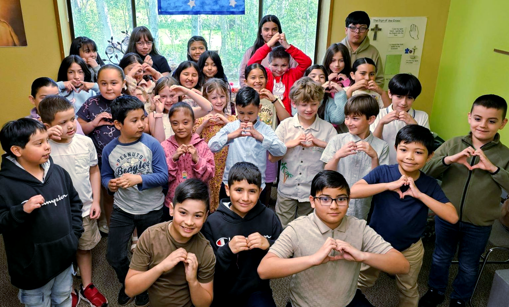 Children posing together indoors, many making hand-heart gestures, with bright classroom walls in the background.