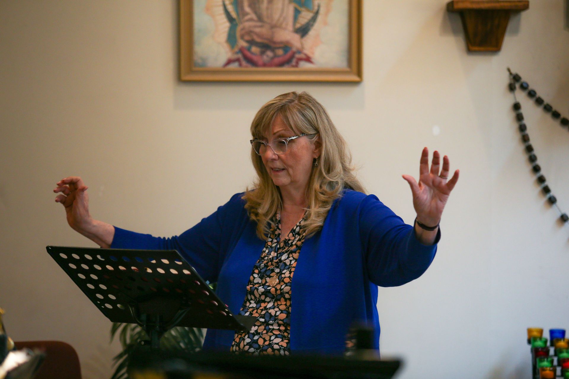 Woman conducting choir with raised hands beside a music stand in a room with wall art.