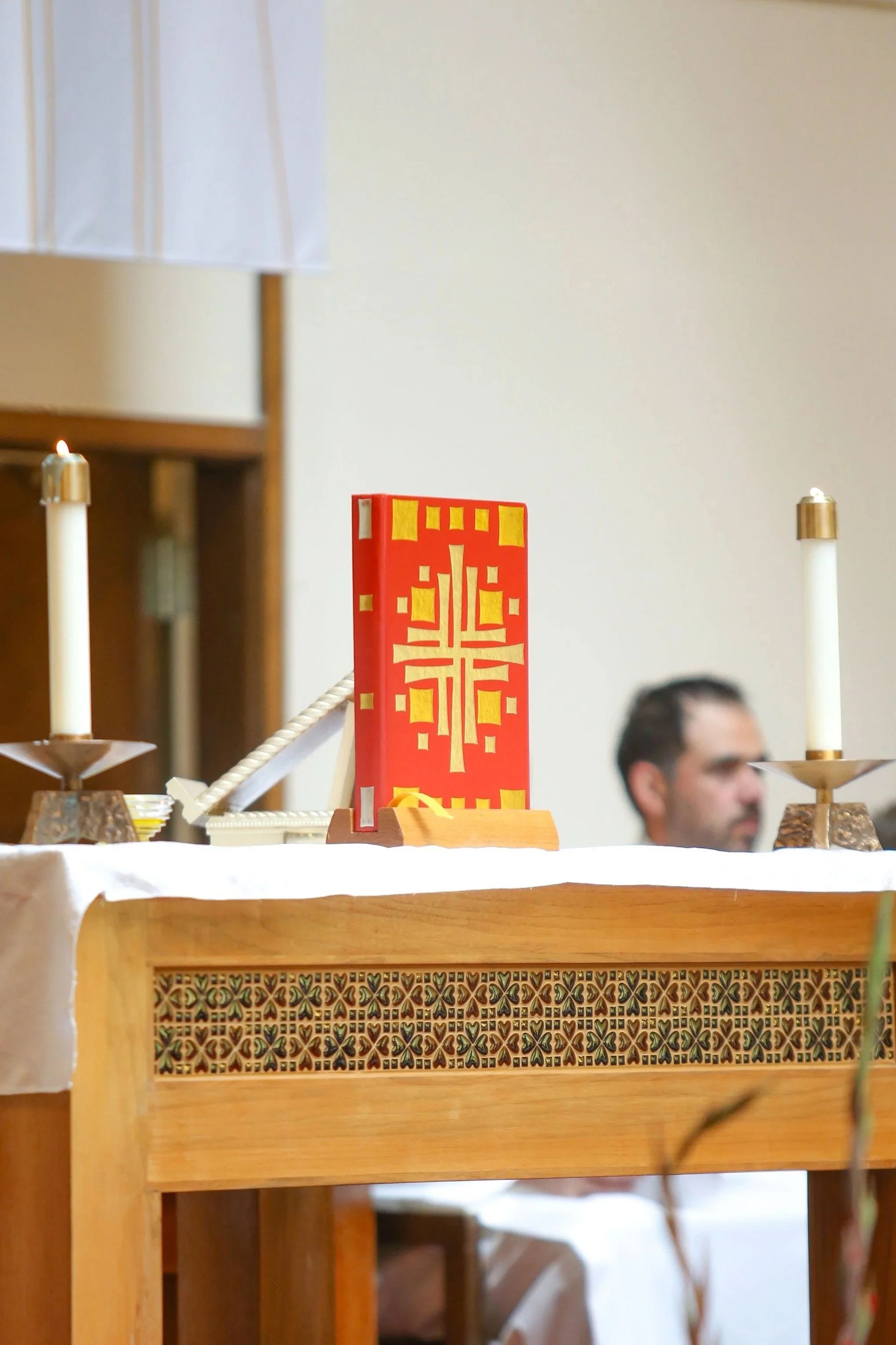 Church altar with a red and gold liturgical book, candles, and a man blurred in the background.