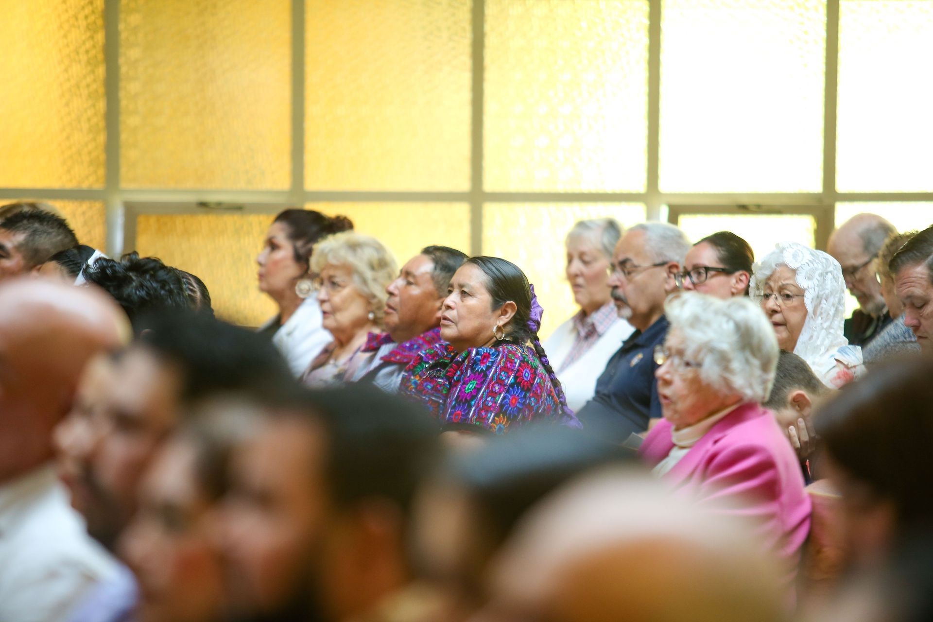 Church people seated in mass, listening attentively, with a yellow-lit windowed wall behind them