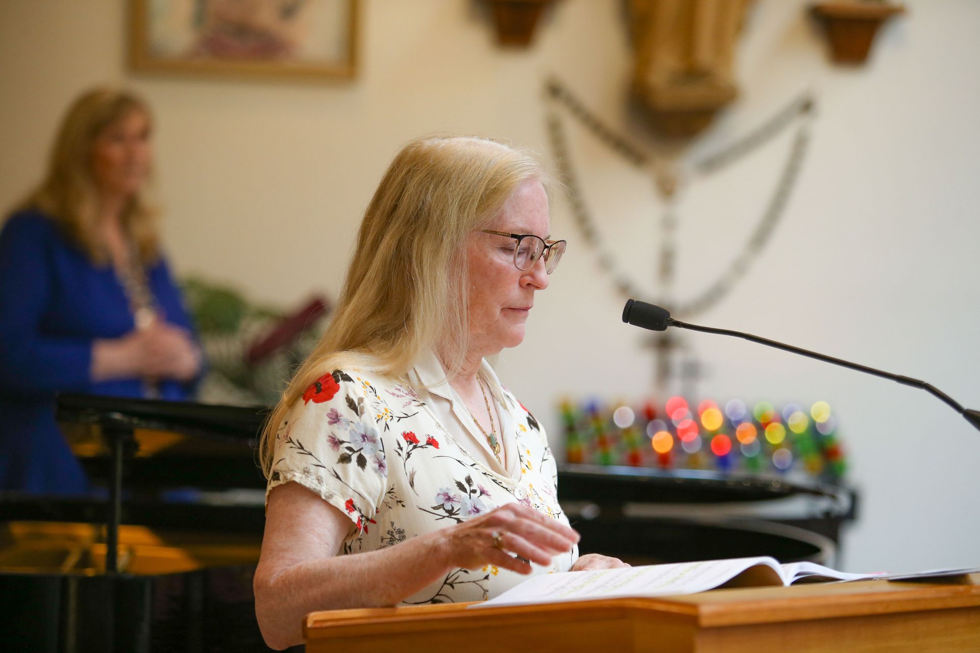 Woman speaking at a podium in a church, with another person blurred in the background.