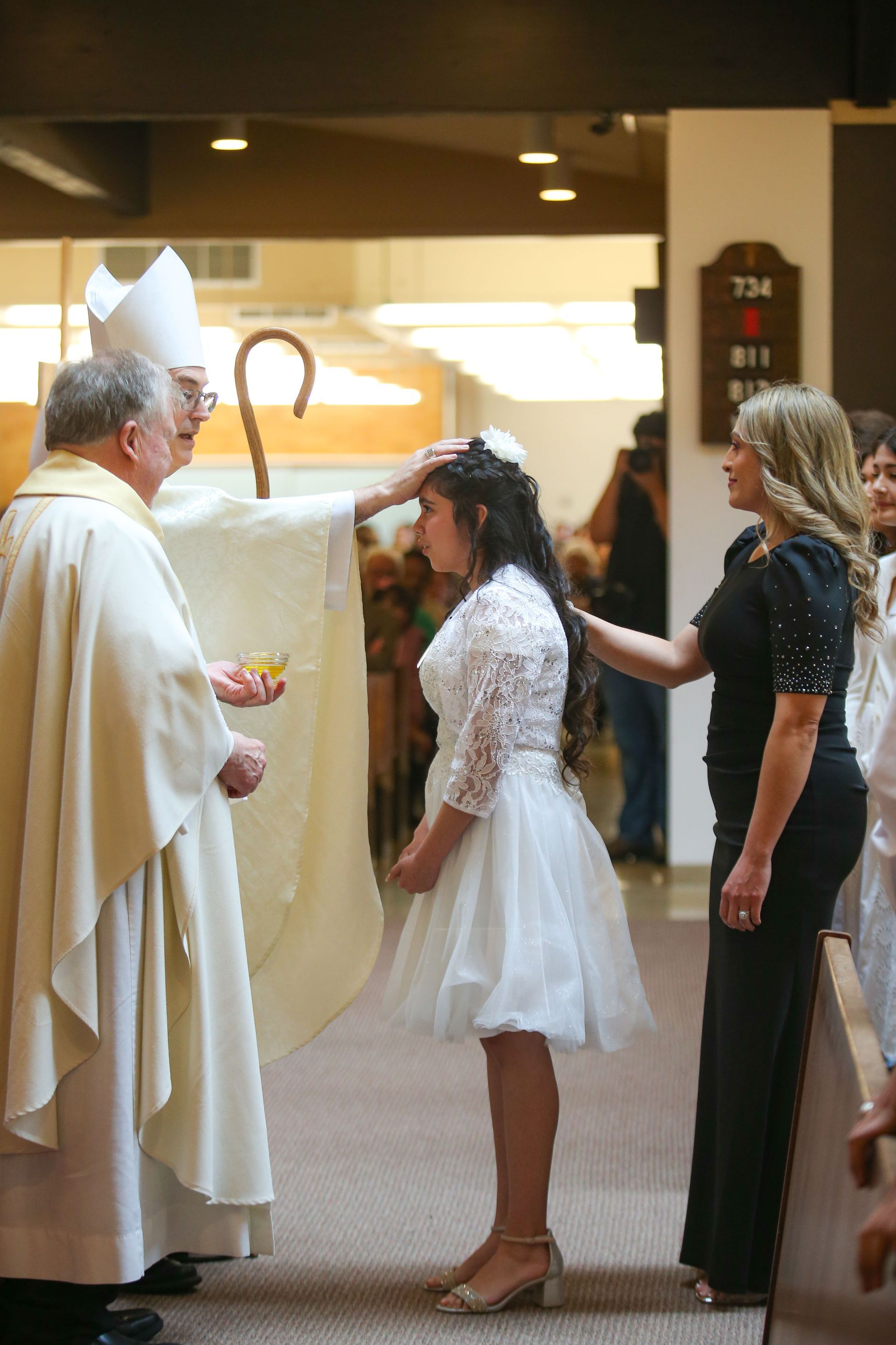 Girl in white dress in front of woman receiving an anointing from the bishop at confirmation mass