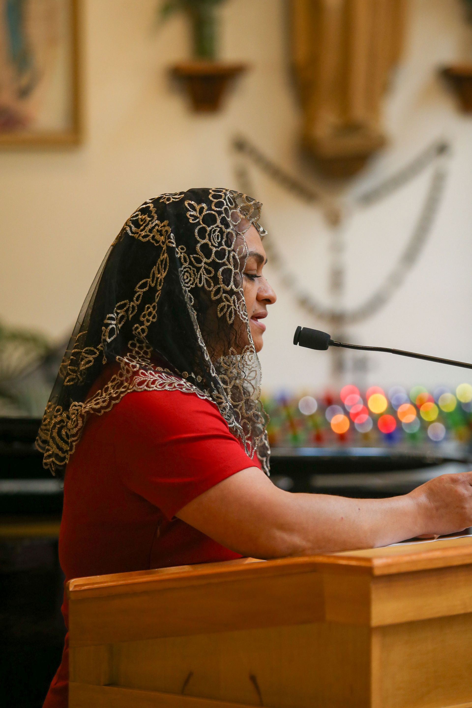 Woman in red speaks at a lectern in a church, wearing a black lace veil.