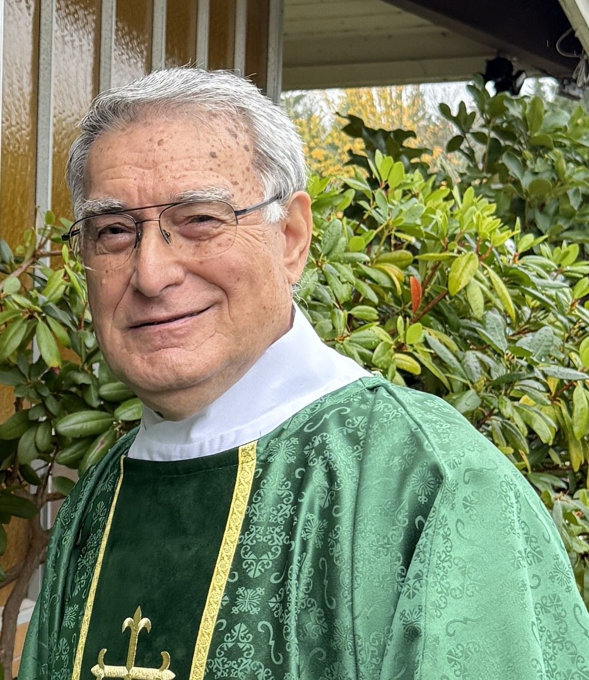 Deacon Henry in green vestments smiling outdoors beside leafy shrubs.