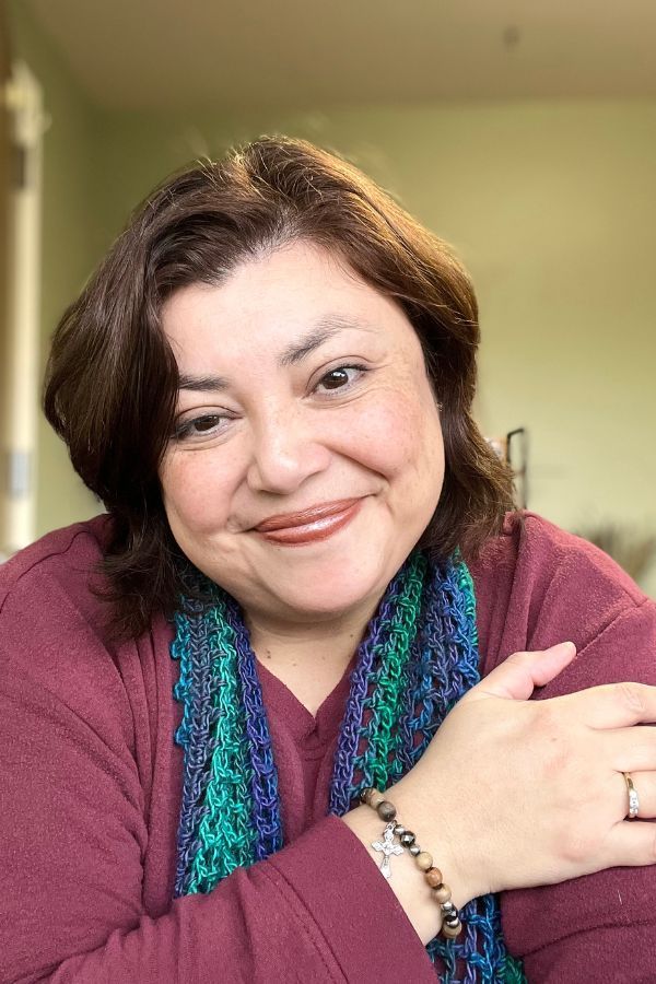 Diana Smiling in a maroon sweater and blue-green scarf, leaning on her hand indoors.