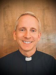 Smiling Fr. Phil in a black clerical shirt with white collar against a warm brown backdrop