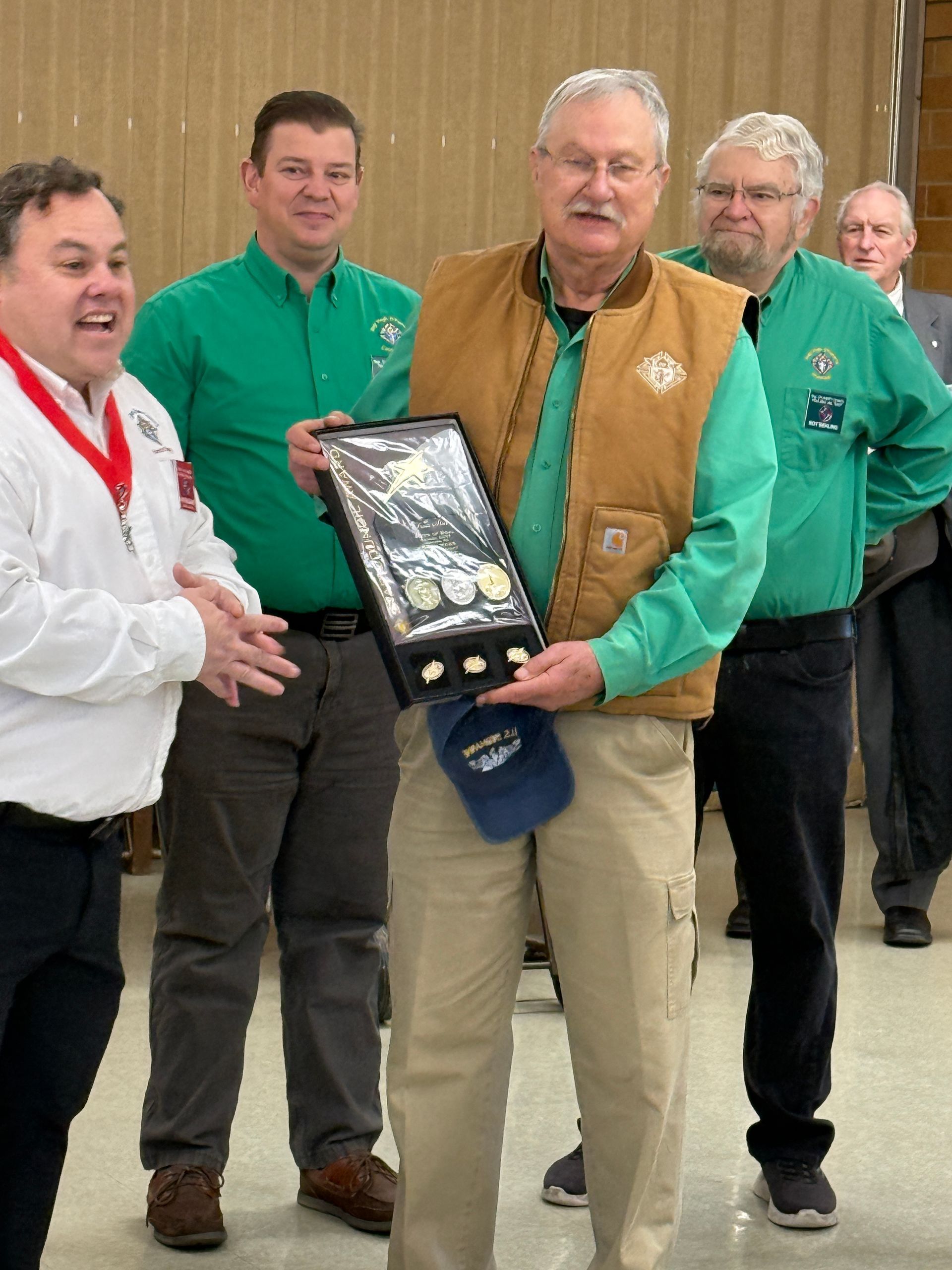 Five men in green shirts hold a framed award indoors, with the man in front smiling.